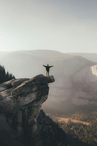 Person with arms raised standing on cliff edge, surrounded by scenic mountain landscape, symbolizing freedom.