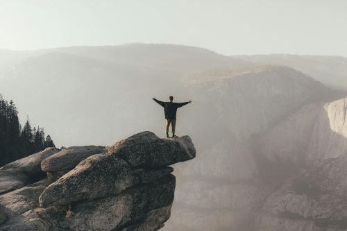 Person with arms raised standing on cliff edge, surrounded by scenic mountain landscape, symbolizing freedom.