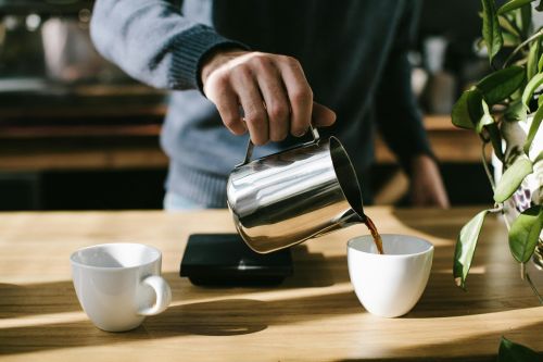 Barista pours steaming hot coffee into a white cup in a cozy indoor setting.