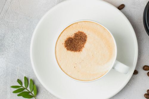 A cappuccino with a heart-shaped cocoa dusting on a saucer, surrounded by coffee beans.
