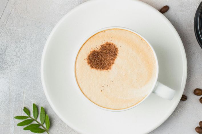 A cappuccino with a heart-shaped cocoa dusting on a saucer, surrounded by coffee beans.