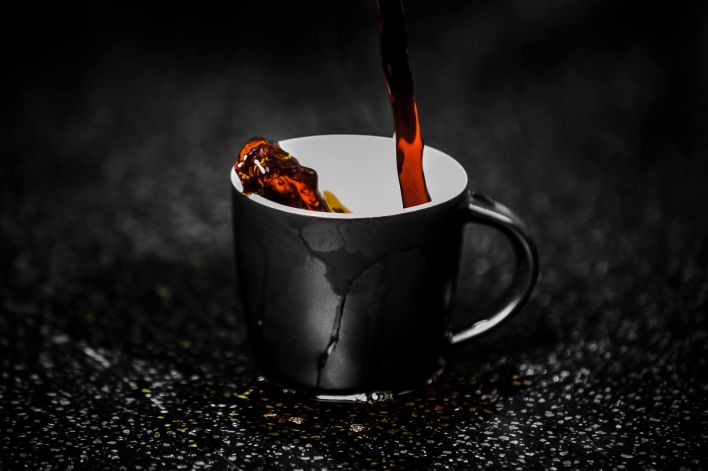 Close-up of steaming dark coffee being poured into a black cup on a dark textured surface. Rich aroma.