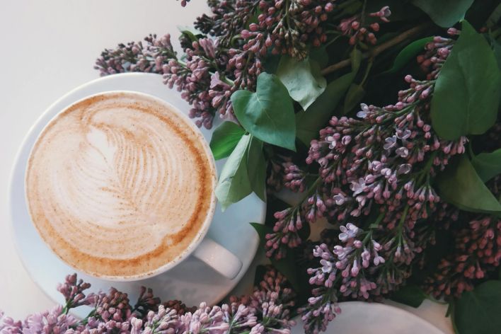 Artistic latte art surrounded by blooming lilac flowers creates a cozy, aesthetic vibe.